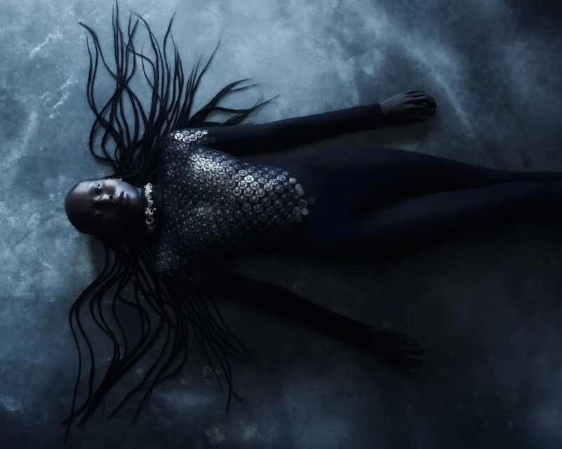 Fashion shot of a dark-skinned model in a dark bodysuit with silver embellishments, laying down with her arms outstretched and hair spread out.