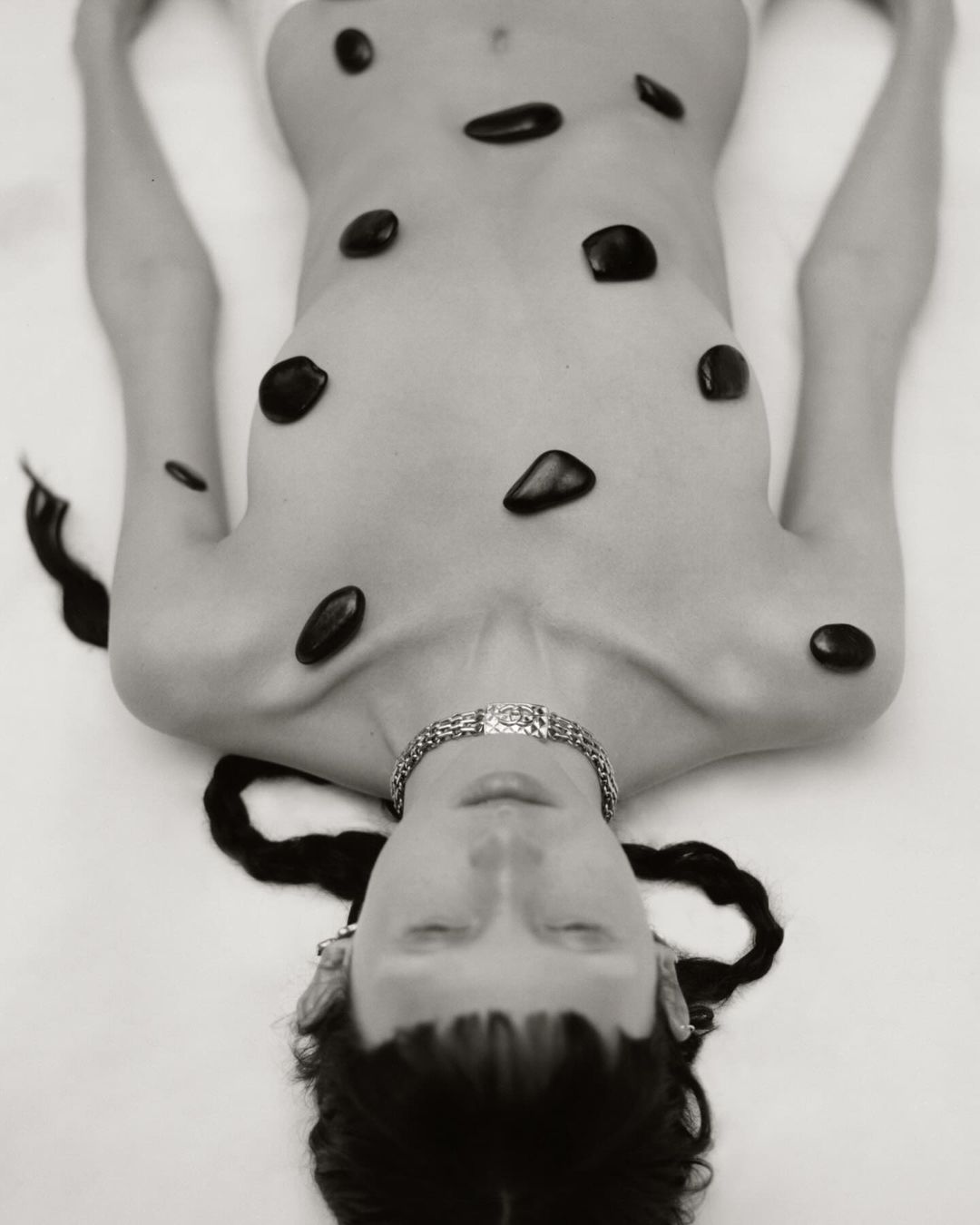 Artistic black and white photo of a woman lying on her back, body adorned with black stones and wearing a silver necklace.