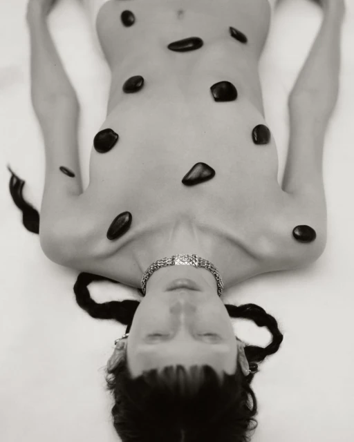 Artistic black and white photo of a woman lying on her back, body adorned with black stones and wearing a silver necklace.