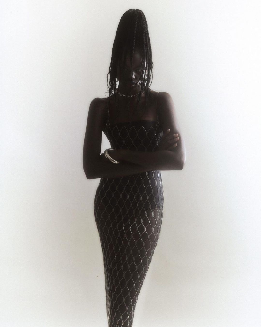 Elegant Black woman in a floor-length gown with a sheer net overlay, posing with arms crossed against a soft white studio backdrop.