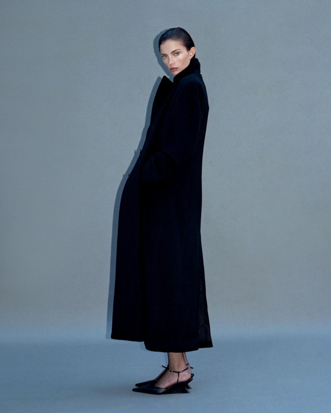 Model in a long black coat and heels standing against a grey backdrop by Myrthe Giesbers