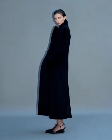 Model in a long black coat and heels standing against a grey backdrop by Myrthe Giesbers