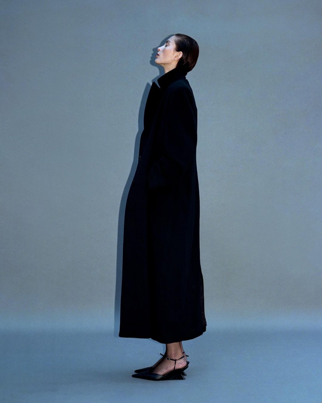 Woman in long black coat and pointed-toe slingbacks posing in profile against a studio backdrop. Fashion editorial photography.