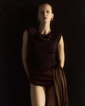 Portrait of a woman wearing a dark top, gold hoop earrings, and a brown draped skirt against a black background.