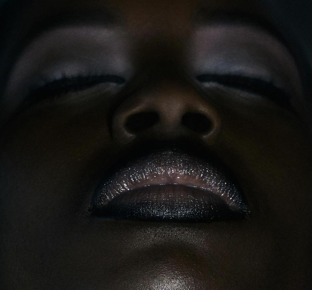 Close-up portrait of an adult female of African descent with closed eyes and dark lipstick in a dark studio environment.