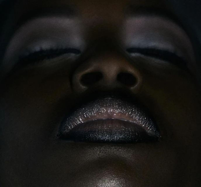 Close-up portrait of an adult female of African descent with closed eyes and dark lipstick in a dark studio environment.