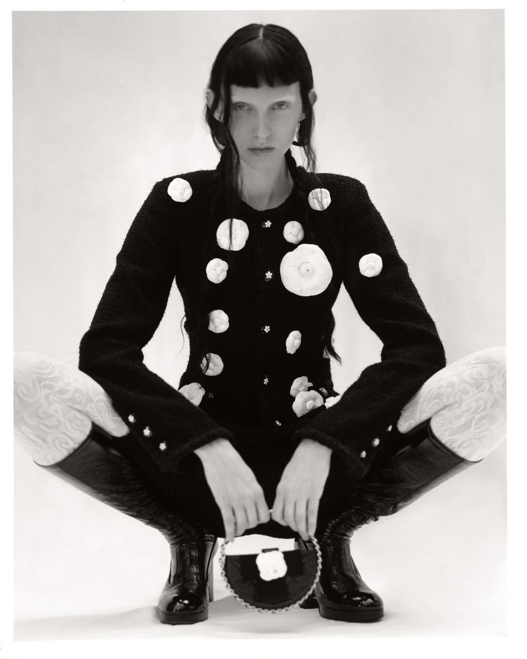 A model with fair skin and dark hair poses in a black and white studio shot, wearing a black jacket and tall boots.
