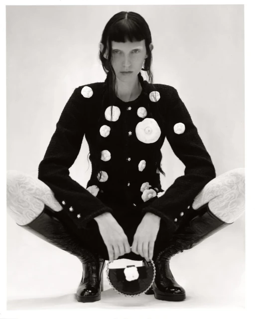 A model with fair skin and dark hair poses in a black and white studio shot, wearing a black jacket and tall boots.