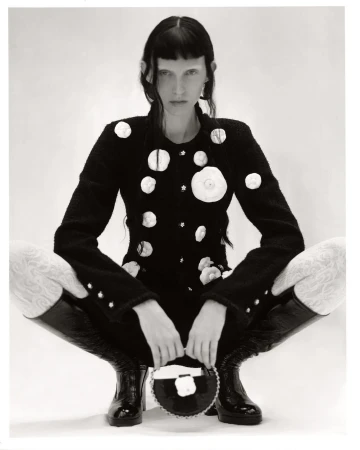 A model with fair skin and dark hair poses in a black and white studio shot, wearing a black jacket and tall boots.