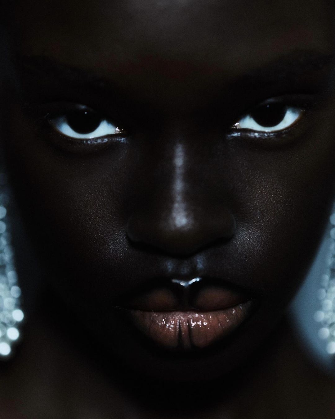 Close-up portrait of a dark-skinned adult female with bright eyes and glossy lips wearing sparkly chandelier earrings in a dark setting.