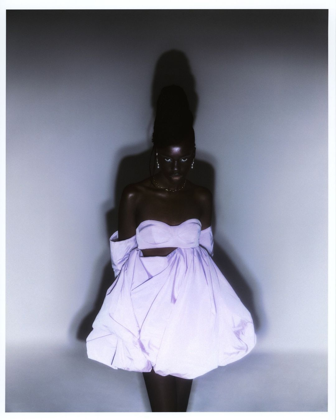 Portrait of dark-skinned adult woman with high ponytail, wearing lavender strapless dress with puffy skirt, in front of grey background.
