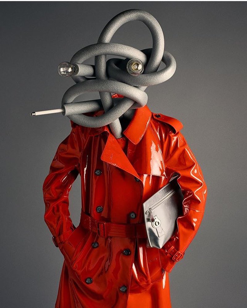 Abstract studio shot of a red vinyl trench coat with a gray foam tubing headpiece and clutch, set against a gray gradient backdrop.
