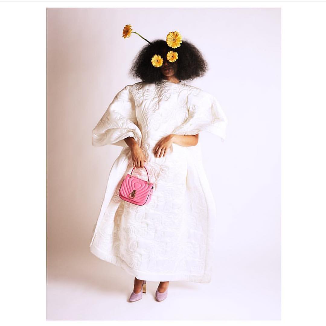 Adult woman wearing puffy white dress and yellow flowers in her afro, holding pink purse, posing in front of a light background.