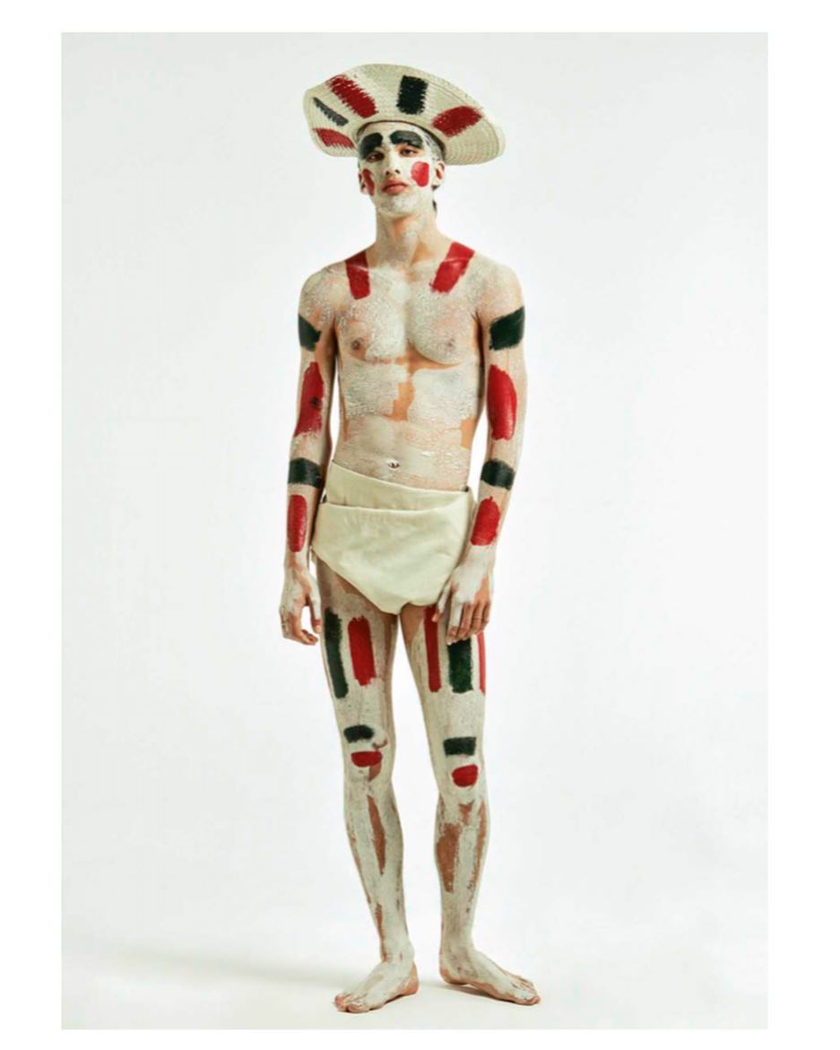 Man with body paint and a wide-brimmed hat poses in a studio. He is shirtless with a cream-colored wrap and paint in red and green.