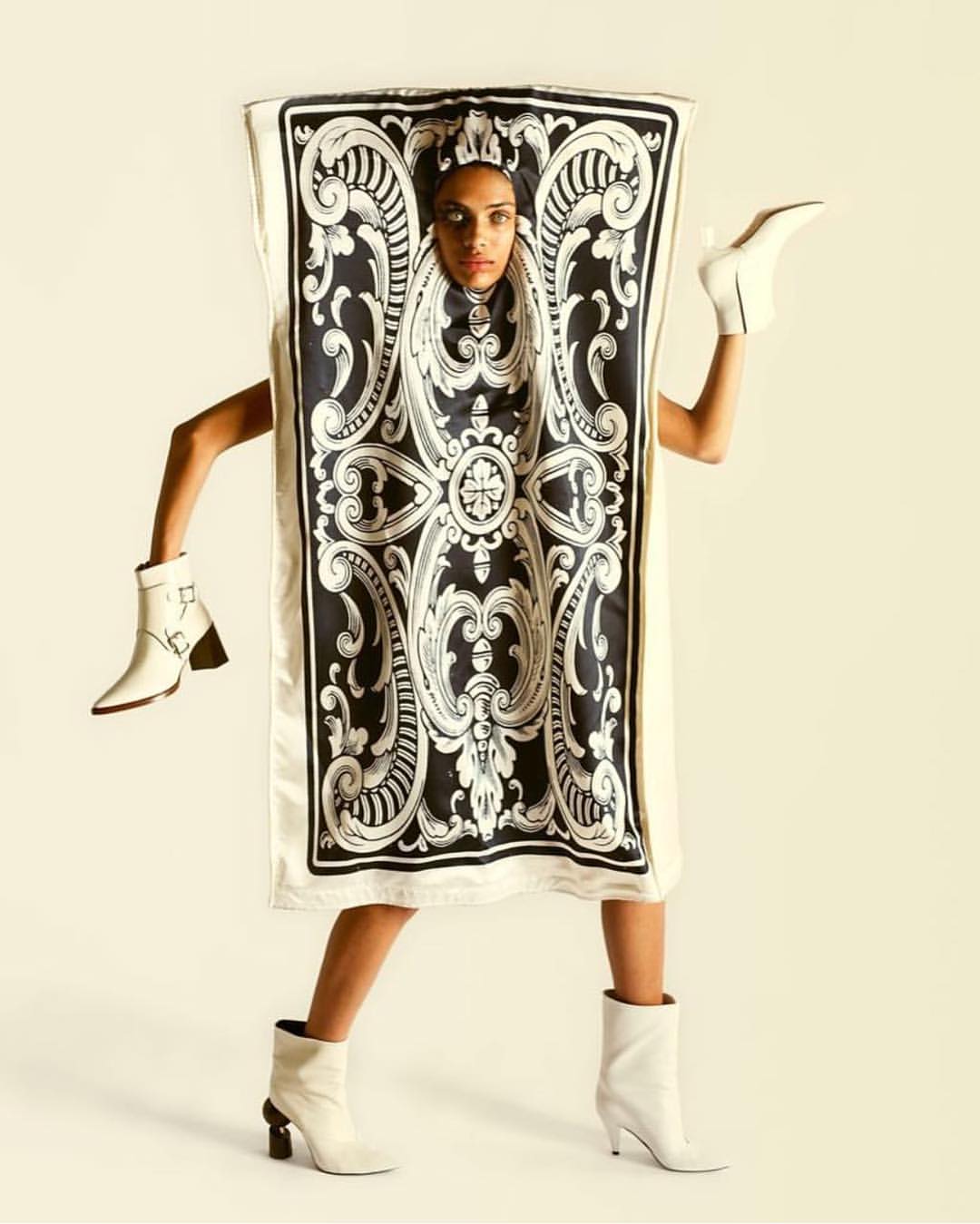Fashion-forward image of a woman posing in a unique garment with black and white patterns, paired with white ankle boots in a studio setting.