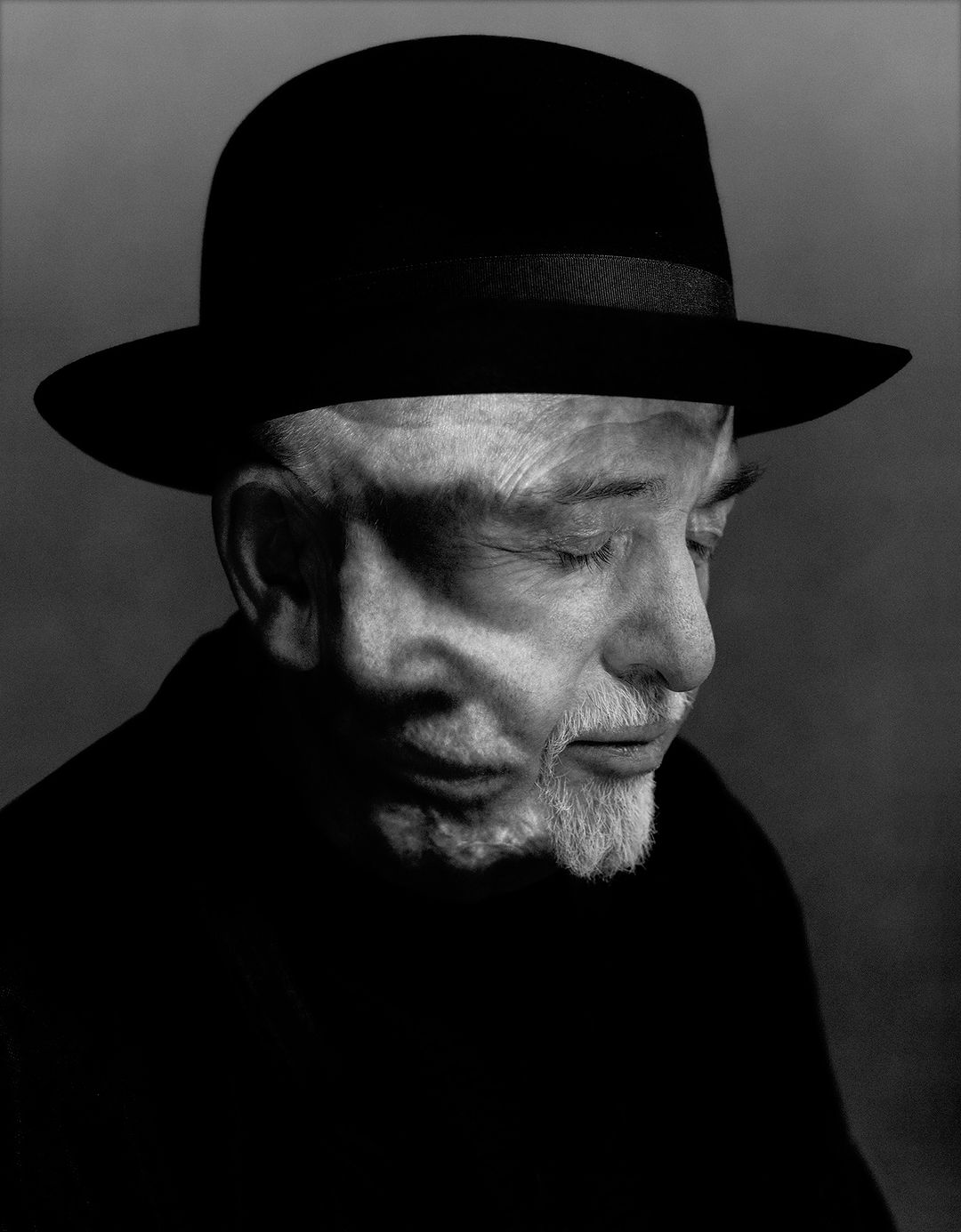 Black-and-white portrait of an elderly man with a white beard wearing a black hat and dark shirt, looking downward with a contemplative expression.