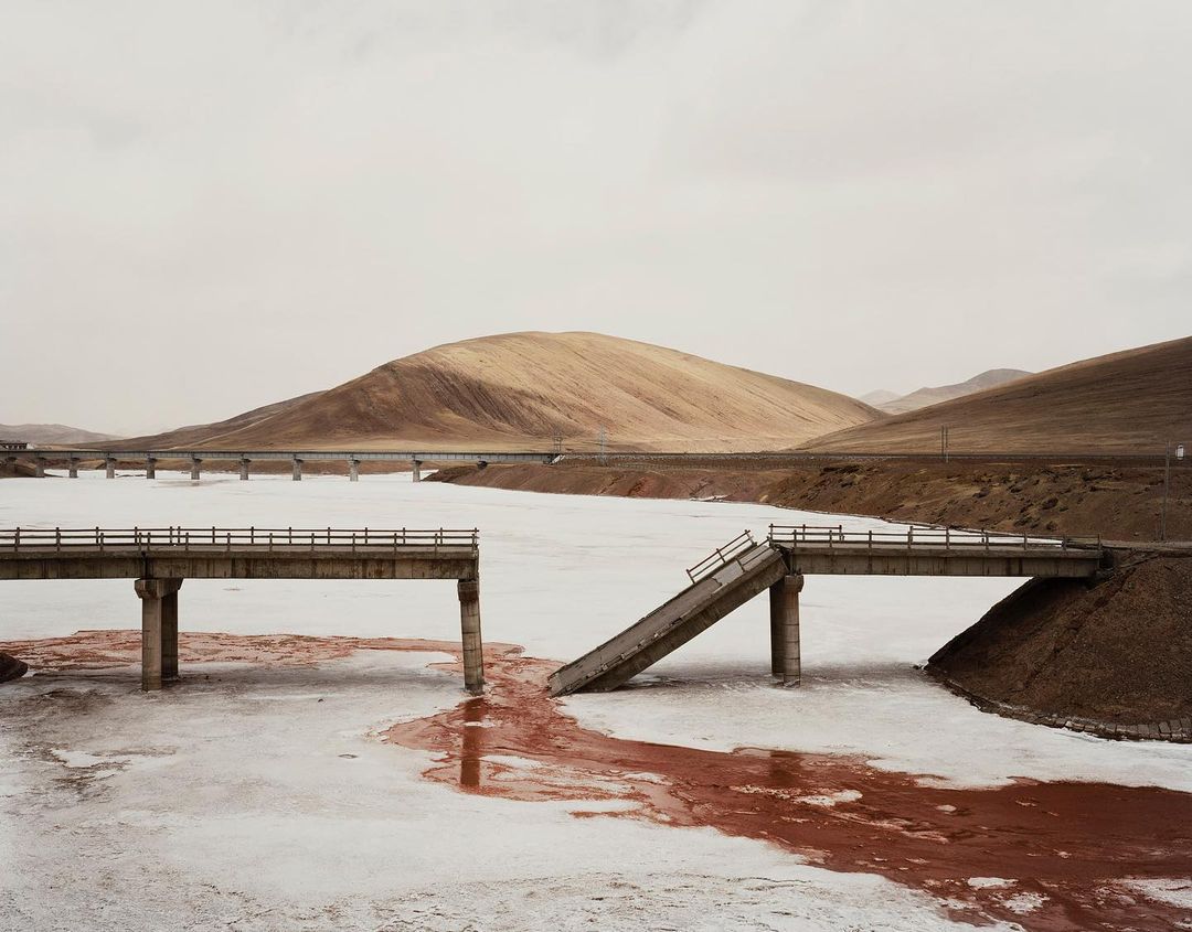 Broken bridge over frozen water with red streaks, leading to a dirt hill in a barren landscape under an overcast sky.