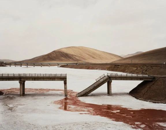 Broken bridge over frozen water with red streaks, leading to a dirt hill in a barren landscape under an overcast sky.