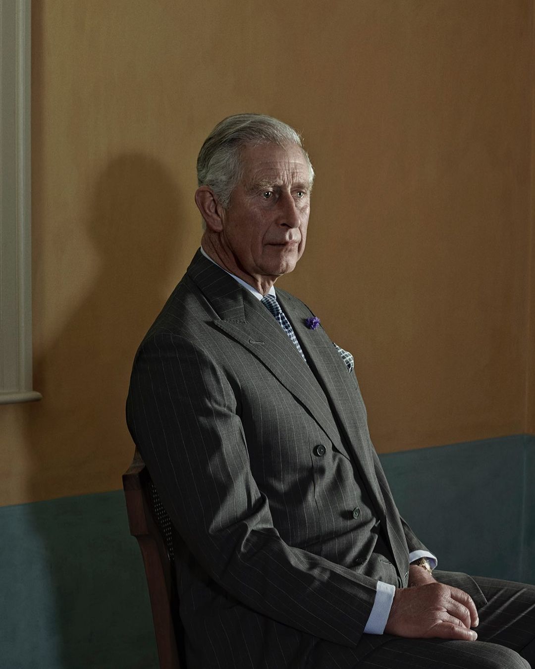 Portrait of an adult male in a grey suit sitting indoors with a tan wall and window in the background, in a studio setting.