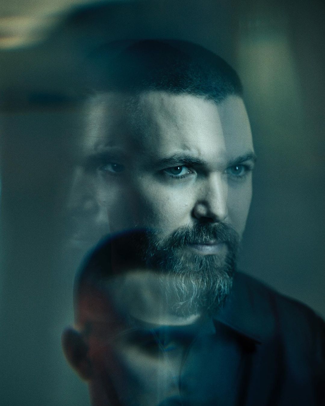 Portrait of a man with a beard with superimposed images, creating a blurred and multi-layered effect in shades of blue and grey.