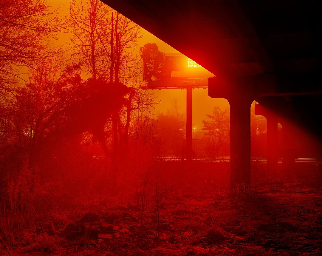 Underneath a bridge with bare trees and a billboard, captured with warm red and orange tones.