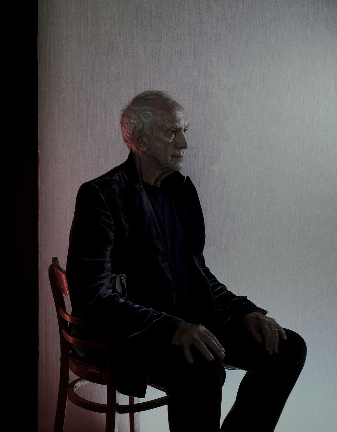 Elderly man in a dark velvet suit sitting on a wooden chair in a photo studio with rim lighting.
