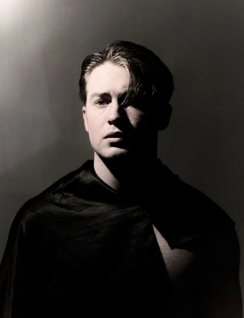 Portrait of a man in deep shadow wearing a dark top under dramatic low key lighting by photographer Nadav Kander.