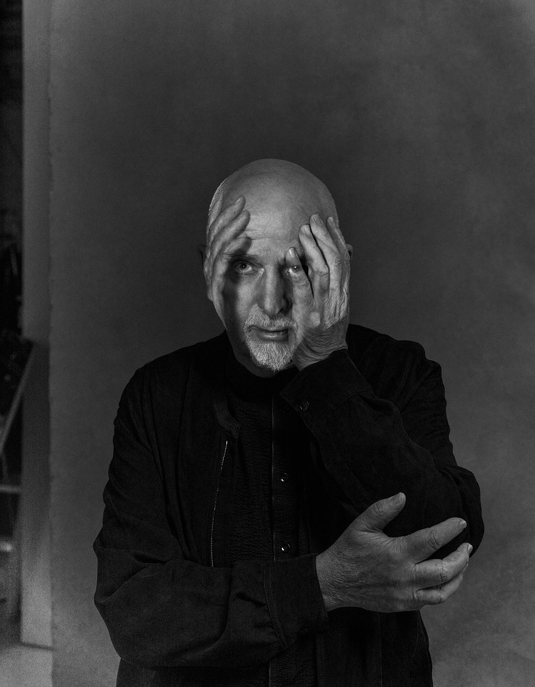 Black and white portrait of a bald man with a short beard, his hands partially covering his face, wearing a black shirt against a dark backdrop.