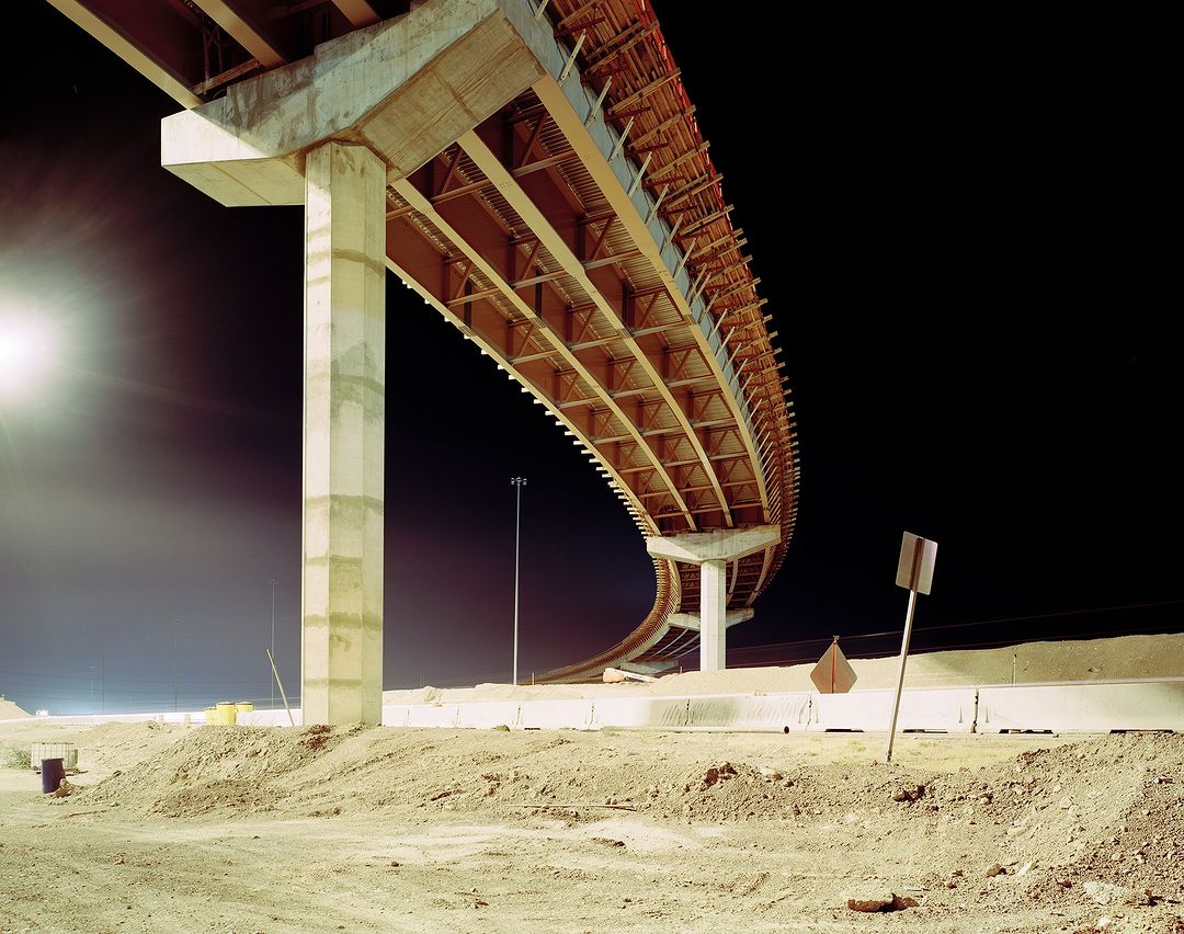 Elevated highway overpass during construction phase, showcasing concrete pillars, wooden support beams, and sandy ground at night.