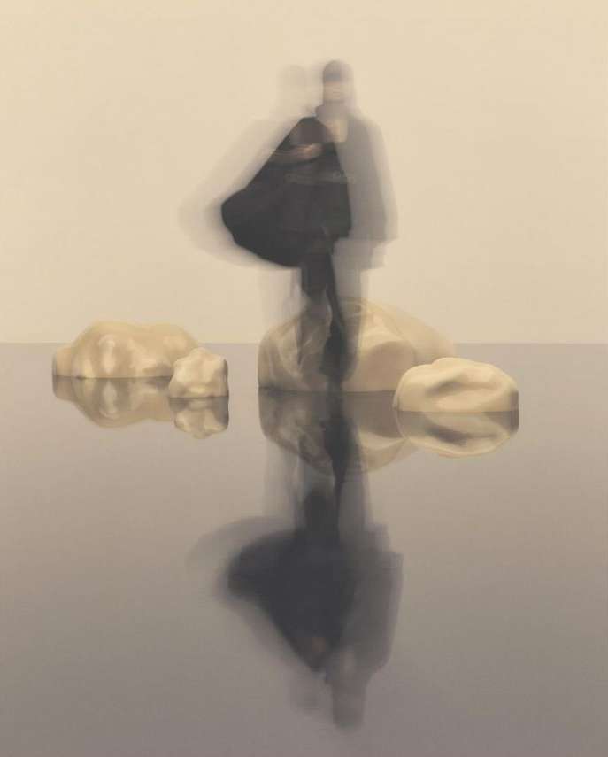 Blurred figures stand on rocks reflected in water against a neutral background, creating a surreal and ethereal photographic artwork.