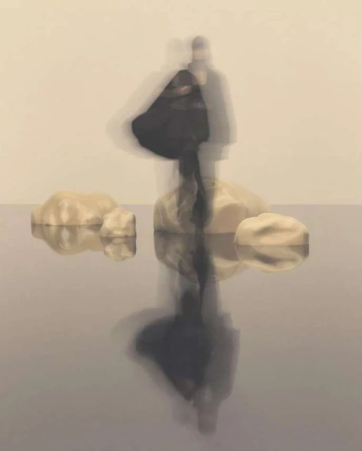Blurred figures stand on rocks reflected in water against a neutral background, creating a surreal and ethereal photographic artwork.