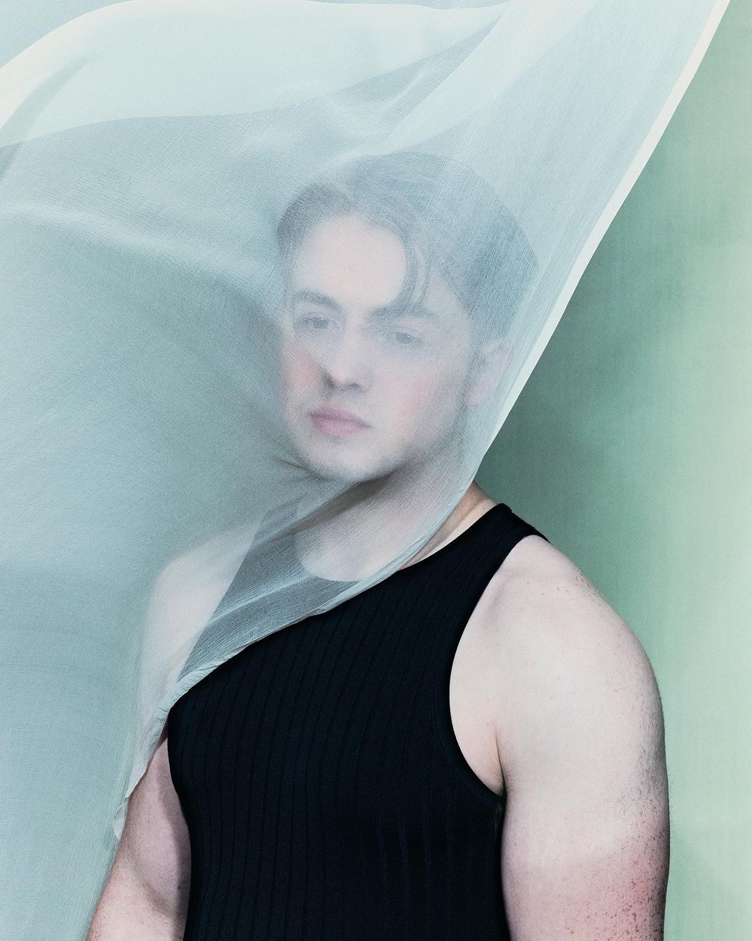 Portrait of a man behind a sheer light green fabric veil wearing a black tank top