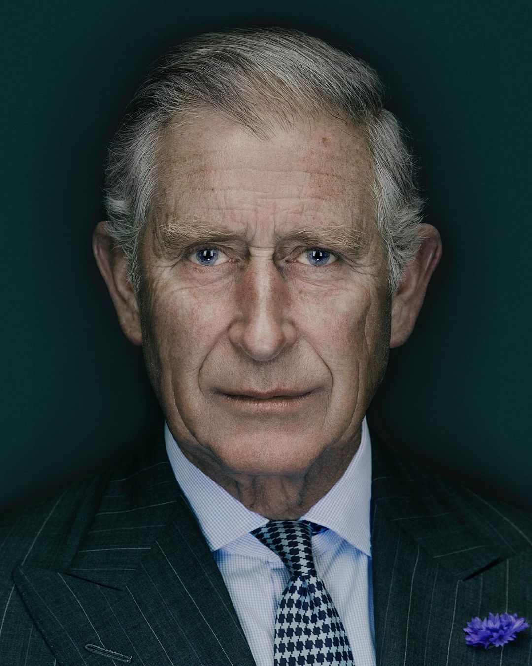 Headshot of an adult male with gray hair wearing a pinstripe suit, light blue shirt, and a patterned tie against a dark teal background.