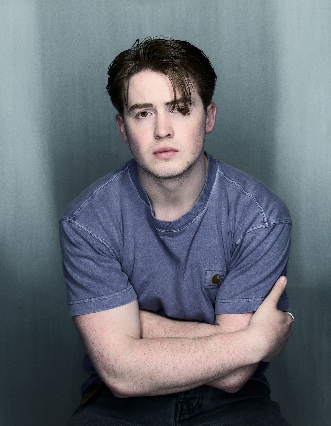 Portrait of a young adult man with brown hair wearing a blue T-shirt, indoors against a grey-blue wall background.