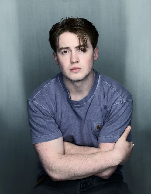 Portrait of a young adult man with brown hair wearing a blue T-shirt, indoors against a grey-blue wall background.