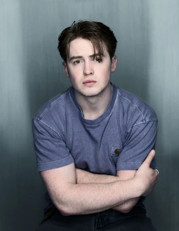 Portrait of a young adult man with brown hair wearing a blue T-shirt, indoors against a grey-blue wall background.