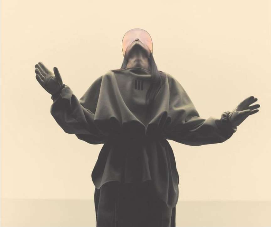 Person in dark hooded garment stands with arms raised against neutral background, with a circular translucent visor.