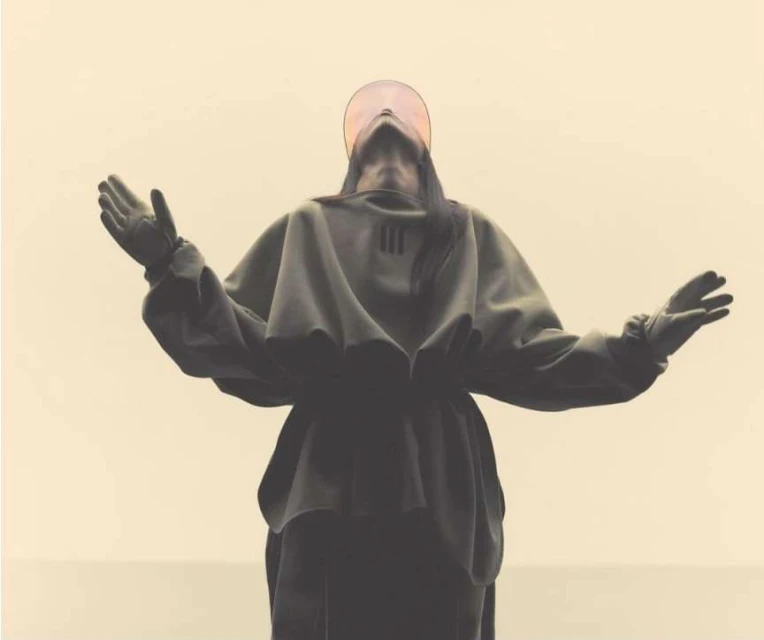 Person in dark hooded garment stands with arms raised against neutral background, with a circular translucent visor.
