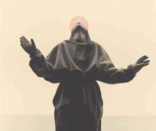 Person in dark hooded garment stands with arms raised against neutral background, with a circular translucent visor.