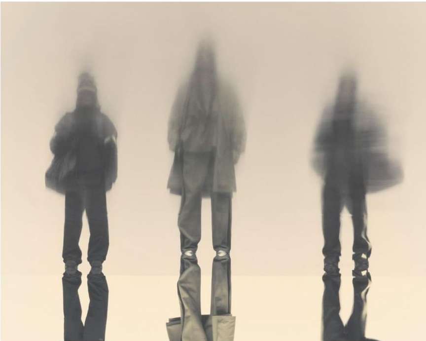 Three blurred figures stand against a light background, silhouetted with undefined features, creating an ethereal and abstract atmosphere.