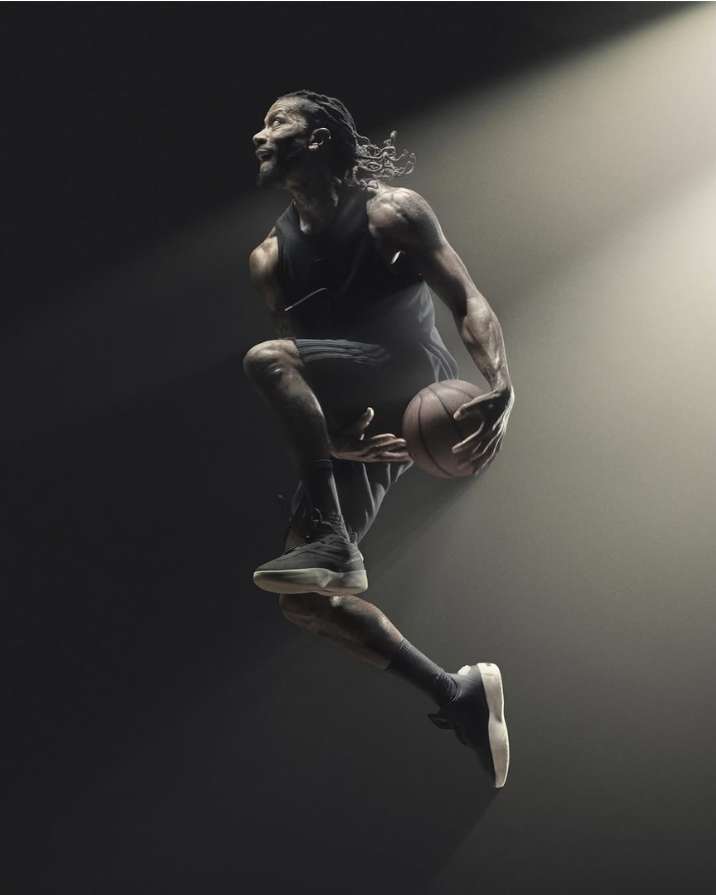 Athletic man in a black tank and shorts mid-air holding a basketball in a dimly lit sports photography setting with dramatic spotlighting.