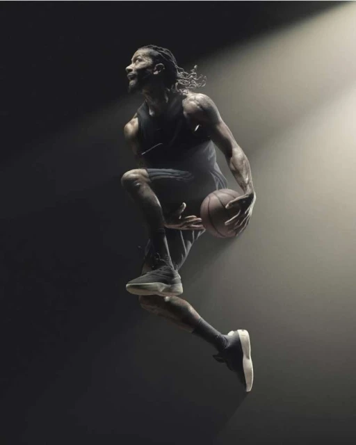 Athletic man in a black tank and shorts mid-air holding a basketball in a dimly lit sports photography setting with dramatic spotlighting.