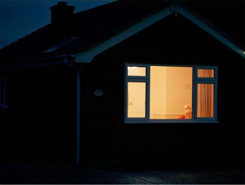 Exterior of a dark brick house at night with a brightly lit window revealing two elderly people sitting inside.