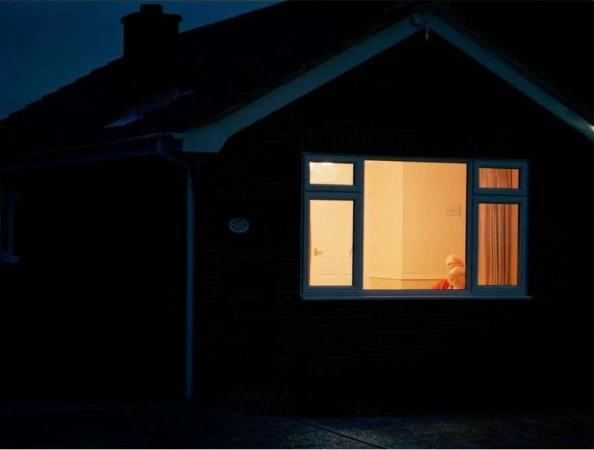 Exterior of a dark brick house at night with a brightly lit window revealing two elderly people sitting inside.