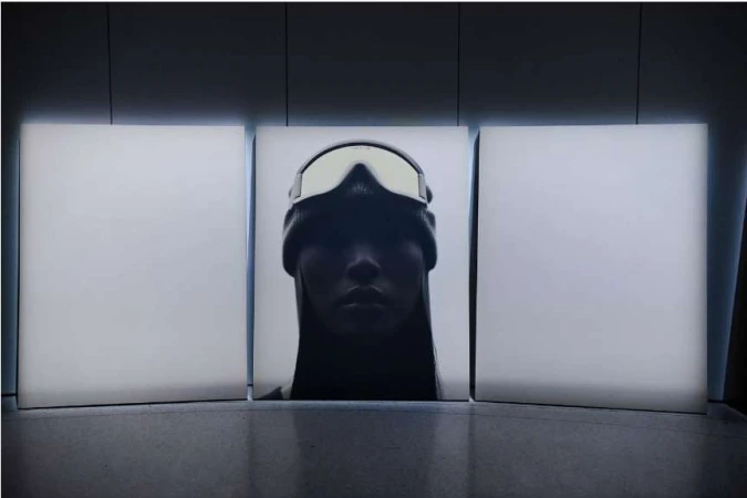 Portrait of a woman in a beanie and ski goggles against three vertically stacked white panels in a dimly lit indoor setting.