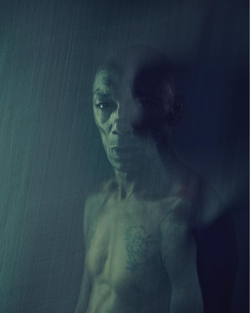 Striking portrait of a shirtless adult man with a tattoo, captured in a dark, textured, abstract, and evocative photo studio setting.