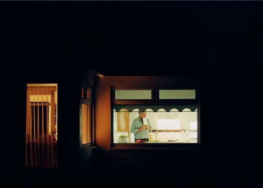 An adult male stands behind a counter in a brightly lit interior, visible through a large window in a darkened exterior setting at night.