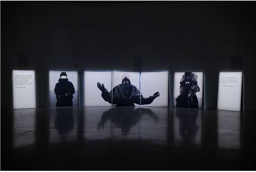 Dark indoor space features images on white panels of people and text, with glossy floor reflections, creating an art installation atmosphere.
