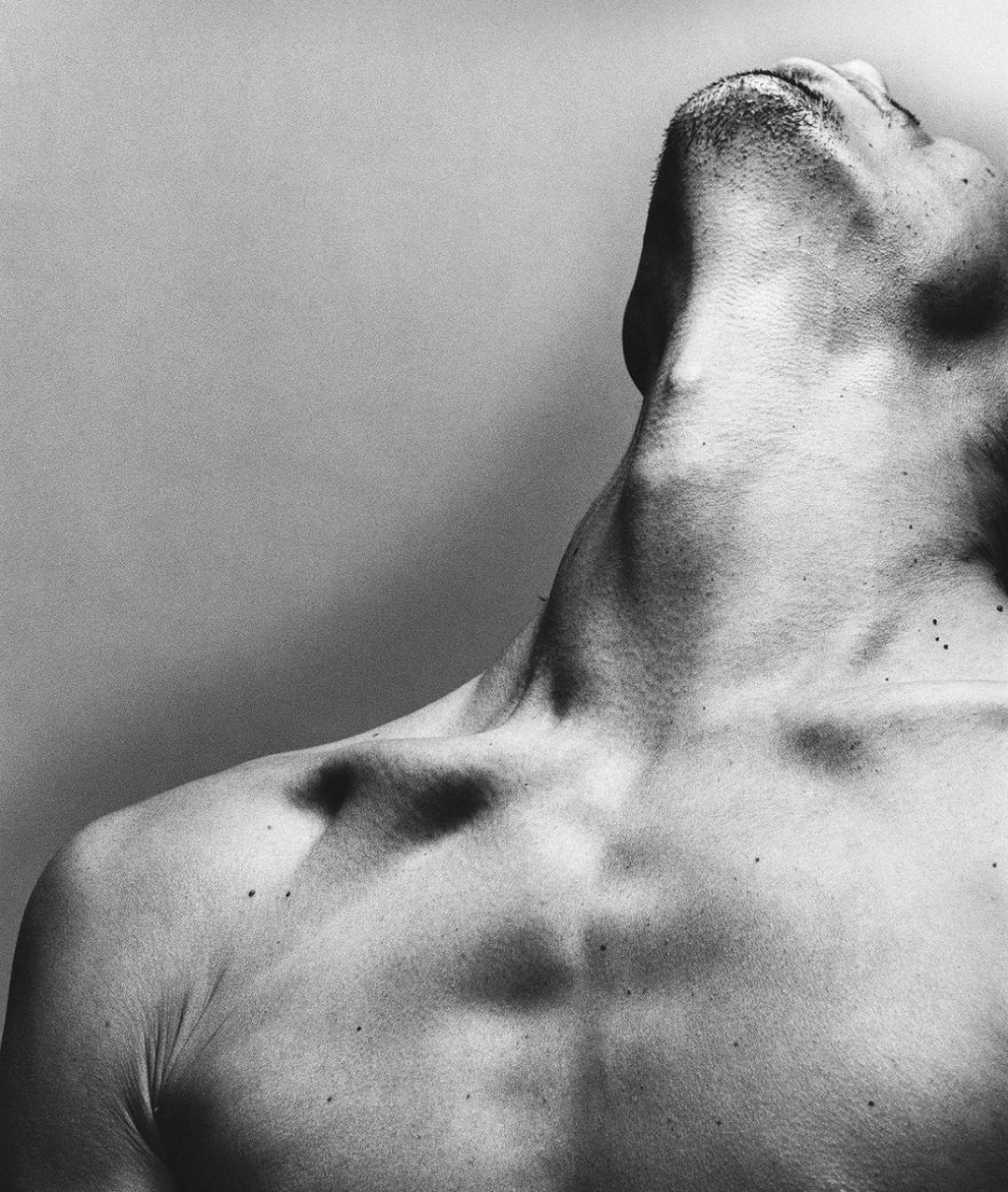 Monochrome portrait of an adult man with his head tilted back, showcasing his neck, chest, and shoulder against a neutral backdrop.