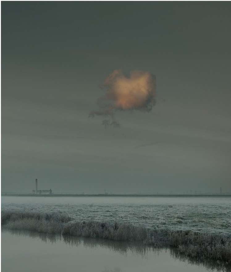 Low-angle landscape captures a cloud illuminated by the sun above a field and calm water, industrial tower in the distant fog.
